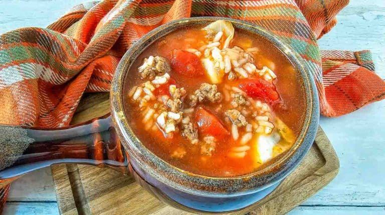 A bowl of crockpot cabbage roll soup with ground beef, rice, tomatoes, and cabbage is placed on a wooden board with an orange plaid cloth nearby.