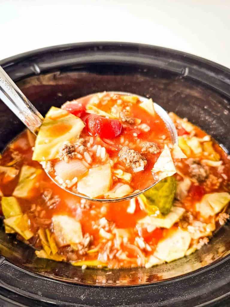 Ladle lifting a serving of cabbage roll soup from a crockpot, showing chunks of cabbage, ground beef, rice, and tomatoes in a tomato-based broth.