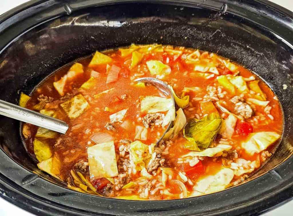 Crockpot filled with cabbage roll soup, showing ground beef, cabbage, rice, and tomatoes in a rich broth with a ladle resting inside.