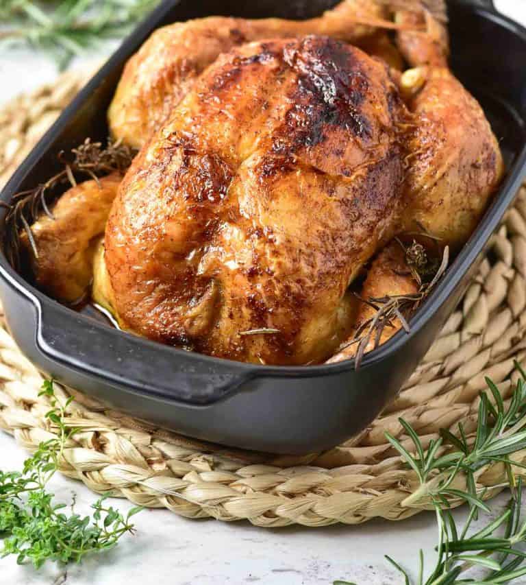 Whole roasted chicken with crispy, golden skin in a black baking dish, garnished with fresh rosemary sprigs.