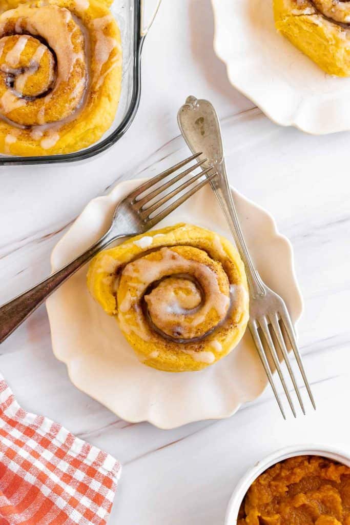 A pumpkin cinnamon roll with icing is shown on a white plate and fork. The background shows more rolls in a baking dish and a small bowl of pumpkin puree.