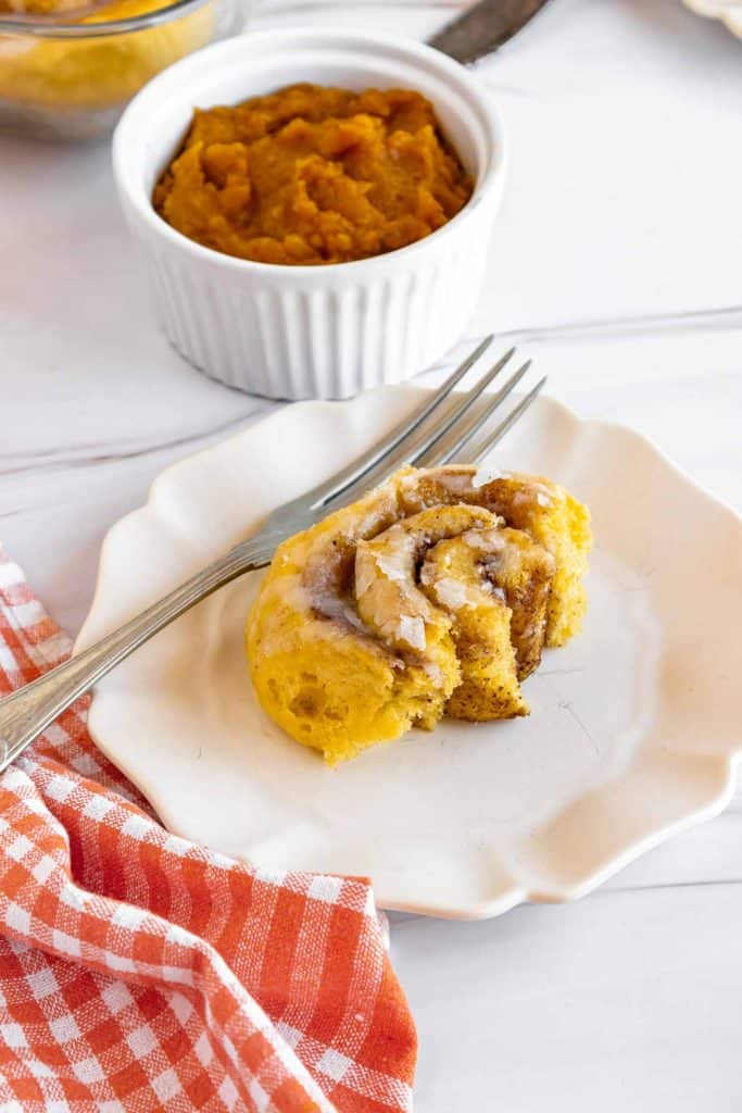 A pumpkin cinnamon roll with icing is shown on a white plate and fork. The background shows more rolls in a baking dish and a small bowl of pumpkin puree.