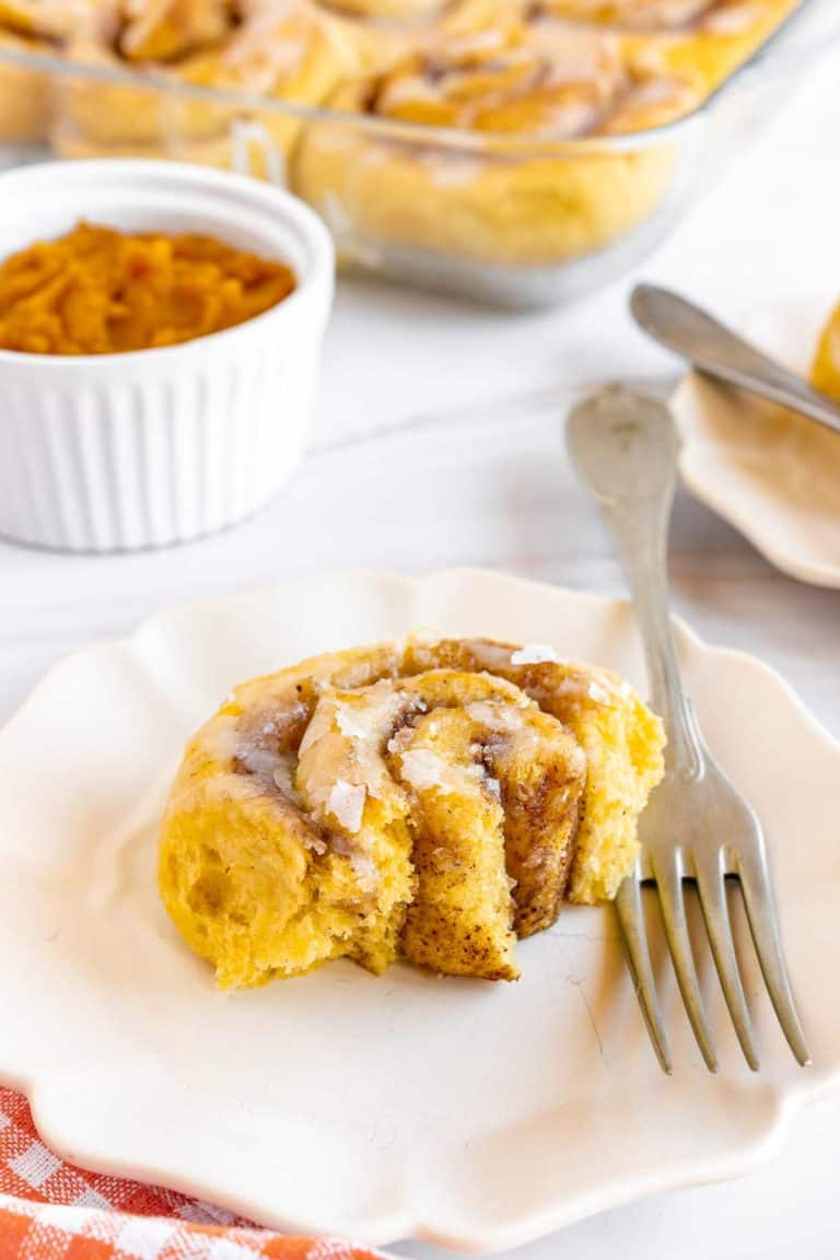 A pumpkin cinnamon roll with icing is shown on a white plate and fork. The background shows more rolls in a baking dish and a small bowl of pumpkin puree.