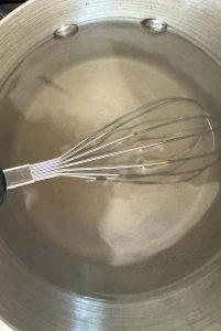 Close-up of a metal whisk in a pot filled with a clear brine solution for pickling.