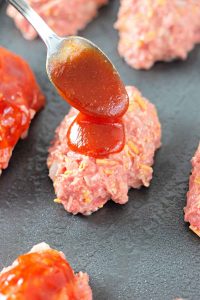 Spoon spreading sauce over mini meatloaves on a baking sheet.