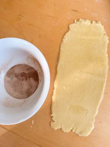 Rolled-out pie crust next to a bowl of cinnamon sugar on a wooden surface.