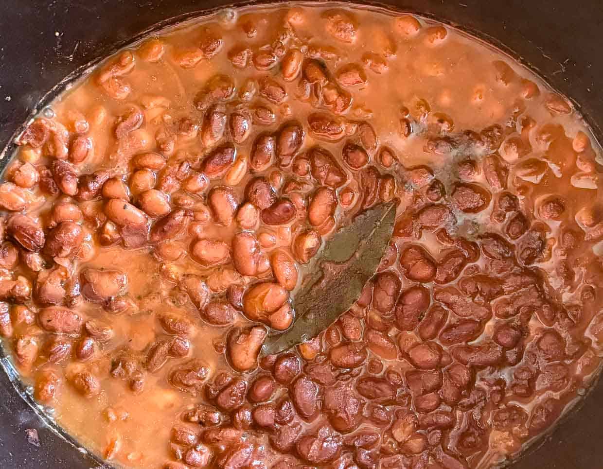 A slow cooker filled with simmering pinto beans in a thick, tomato-based sauce with a visible spoon.