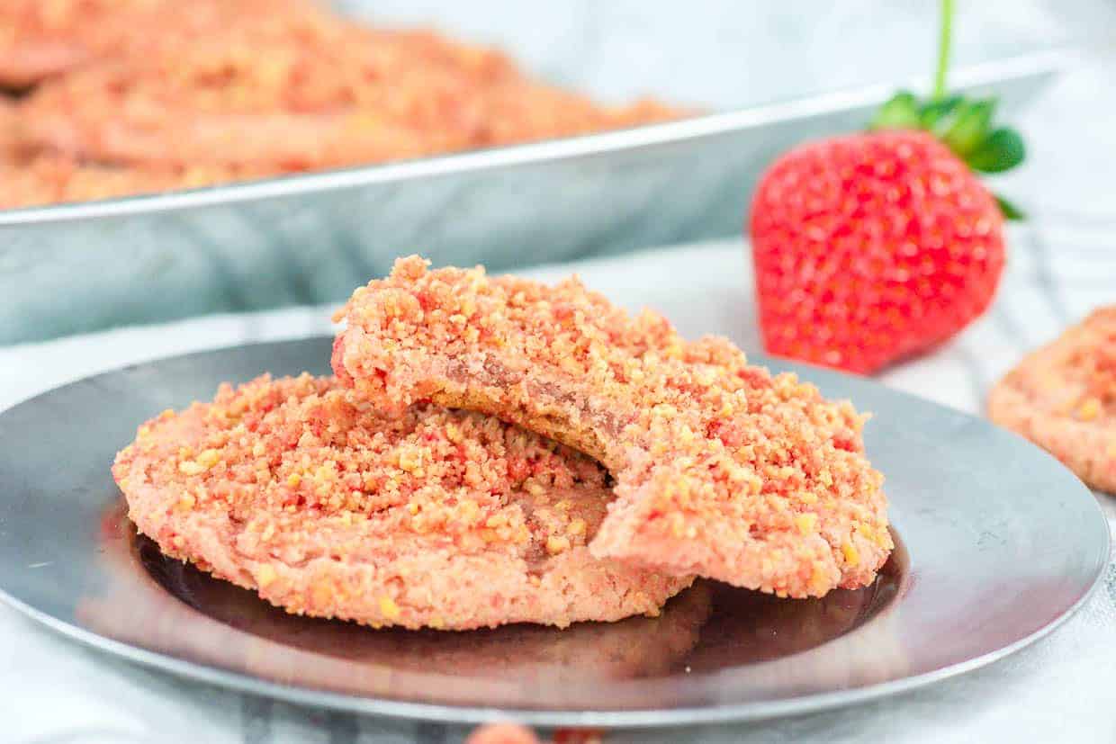 A close-up of Strawberry Crunch Cookies on a reflective metal plate, accompanied by a fresh strawberry.