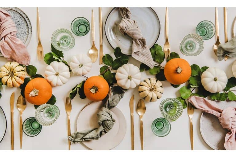 Elegant fall tablescape with mini pumpkins, greenery, gold cutlery, and cloth napkins on plates, arranged with green glassware.