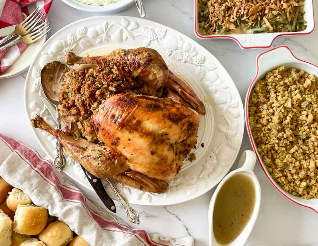 Oven-roasted turkey with stuffing on a platter, served with gravy, green bean casserole, extra stuffing, and dinner rolls.