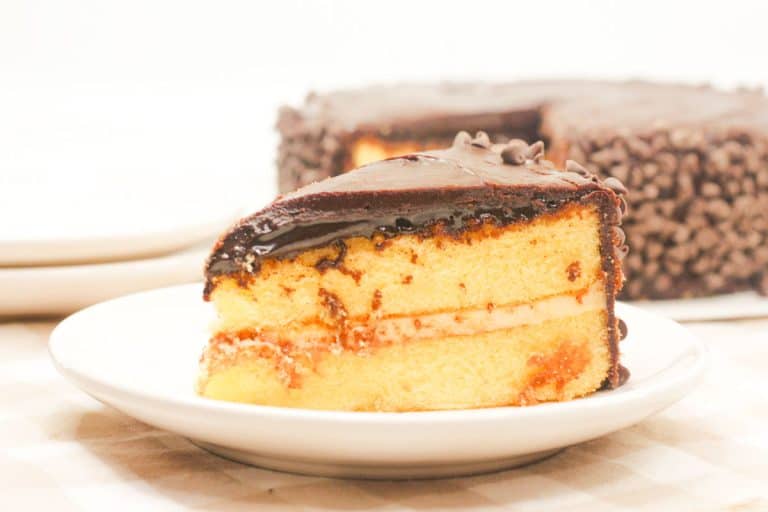 A close-up of a slice of Boston Cream Pie Cake on a white plate, with layers of vanilla cake, custard, and chocolate ganache.
