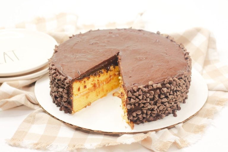 Boston Cream Pie Cake on a white plate, topped with chocolate ganache and coated with chocolate chips on the sides.