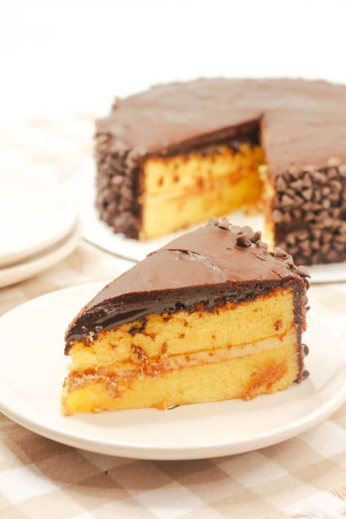 A slice of Boston Cream Pie Cake on a white plate, with the full cake in the background.