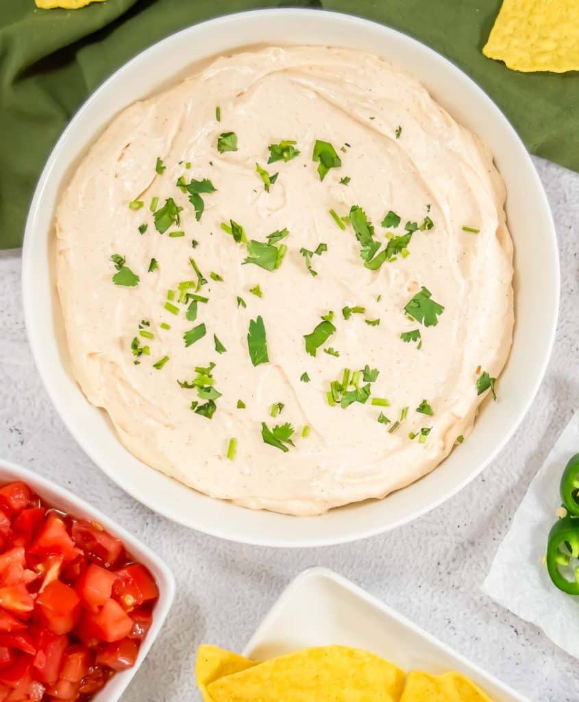 Easy taco dip in a white bowl, topped with cilantro and green onions, with tortilla chips, diced tomatoes, and jalape&ntilde;os around it.