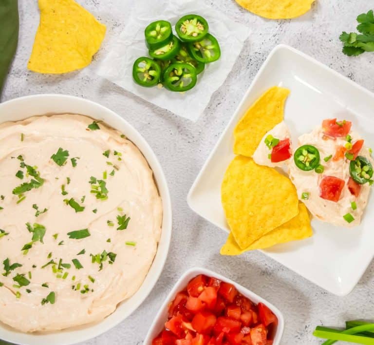 A bowl of creamy dip topped with chopped herbs, surrounded by diced tomatoes, tortilla chips, and sliced jalapeños.