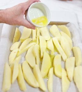 Raw potato wedges on a baking sheet, with melted butter being poured over them.