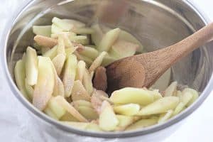 Sliced apples mixed with sugar and cinnamon in a metal bowl with a wooden spoon.