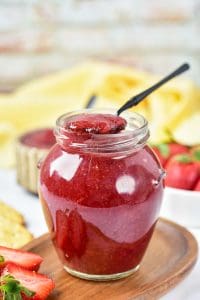 Jar of Easy Strawberry Jam with a spoon, surrounded by fresh strawberries.