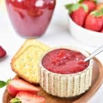 Strawberry jam in a bowl with crackers and halved strawberries on a wooden serving tray.