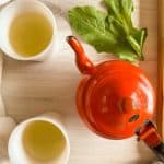 Cups of fresh dandelion greens tea next to an orange teapot and fresh leaves.