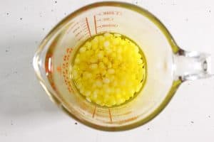 Beeswax granules and dandelion-infused oil combined in a glass measuring cup.
