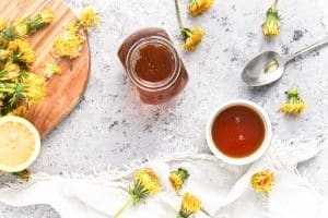 Finished dandelion syrup in a jar and small bowl with fresh dandelions and lemon on the side.