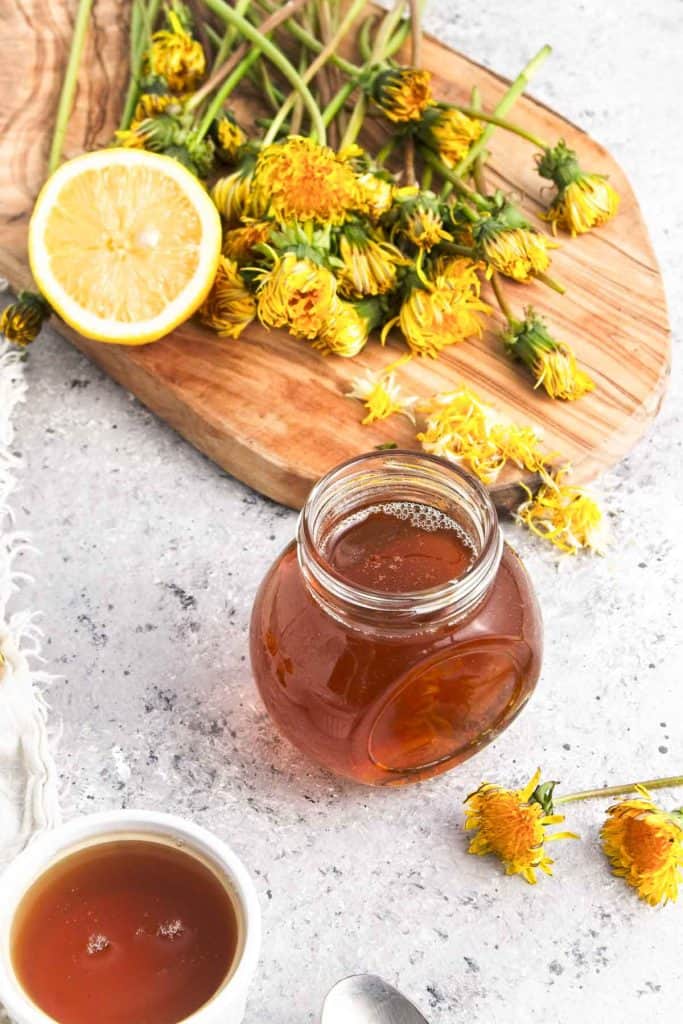 Jar of dandelion syrup with dandelion flowers and half a lemon on a board in the background.