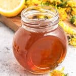 Close-up of a jar of dandelion syrup with fresh dandelions and a lemon on a wooden board behind it.