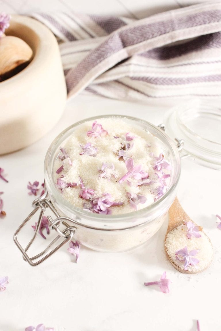Wooden spoon with lilac sugar and lilac blossoms, jar of lilac sugar in the background.