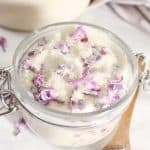 Glass jar filled with granulated sugar and lilac blossoms, surrounded by scattered lilac petals.