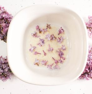 White bowl with granulated sugar and small lilac blossoms floating on top.