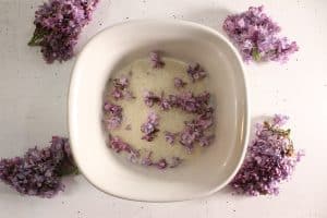 White bowl filled with granulated sugar layered with lilac petals, lilac flowers placed around the bowl.