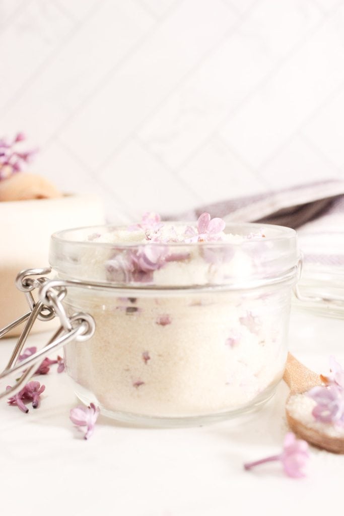 Glass jar filled with granulated sugar and lilac blossoms, surrounded by scattered lilac petals.