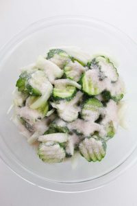 Sliced cucumbers and onions mixed with salt in a glass bowl.