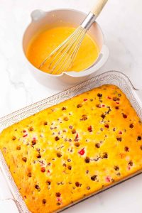 Baked poke cake with holes across the top next to a bowl of orange-colored pudding mixture.