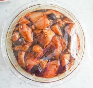 Bowl of raw chicken chunks marinating in teriyaki sauce, covered with plastic wrap.
