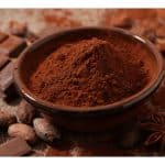 Bowl of cocoa powder surrounded by cocoa beans, chocolate pieces, cinnamon sticks, and star anise on a rustic wooden surface.