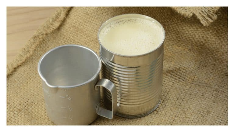 Open can of evaporated milk next to a metal measuring cup.
