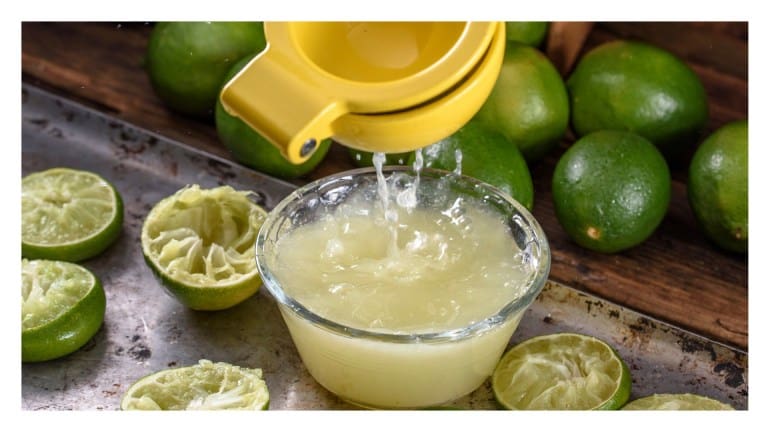 Fresh lime juice being squeezed into a glass bowl with a yellow juicer, surrounded by whole and spent lime halves.