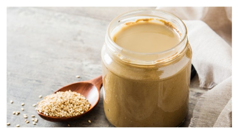 Jar of tahini with smooth surface next to a wooden spoon filled with sesame seeds on a gray background.