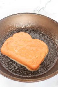 Coated bread slice cooking in a frying pan.
