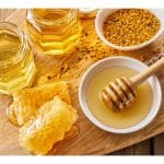 Jars of honey with honeycomb, pollen, and a honey dipper on a wooden board.