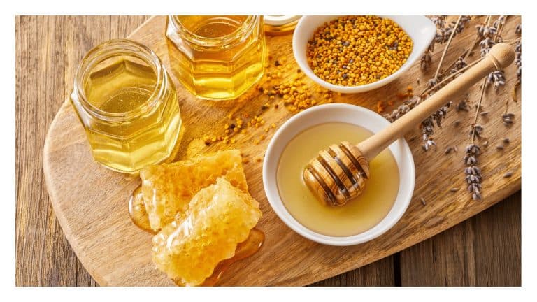 Jars of honey with honeycomb, pollen, and a honey dipper on a wooden board.
