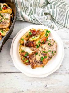 Plated pork chop with roasted apples, onions, and bacon, garnished with parsley.