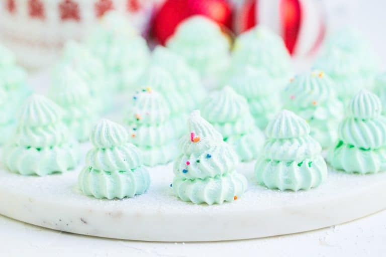 Rows of Christmas tree meringues on a white marble tray with cocoa and ornaments behind them.