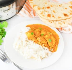 Plate of butter chicken and rice with naan bread and a slow cooker in the background.