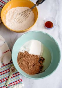 Cocoa, sugar, and oil combined in a teal bowl with flour mixture nearby.