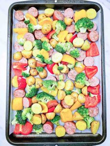 Uncooked sausage, potatoes, broccoli, and peppers arranged on a baking sheet.