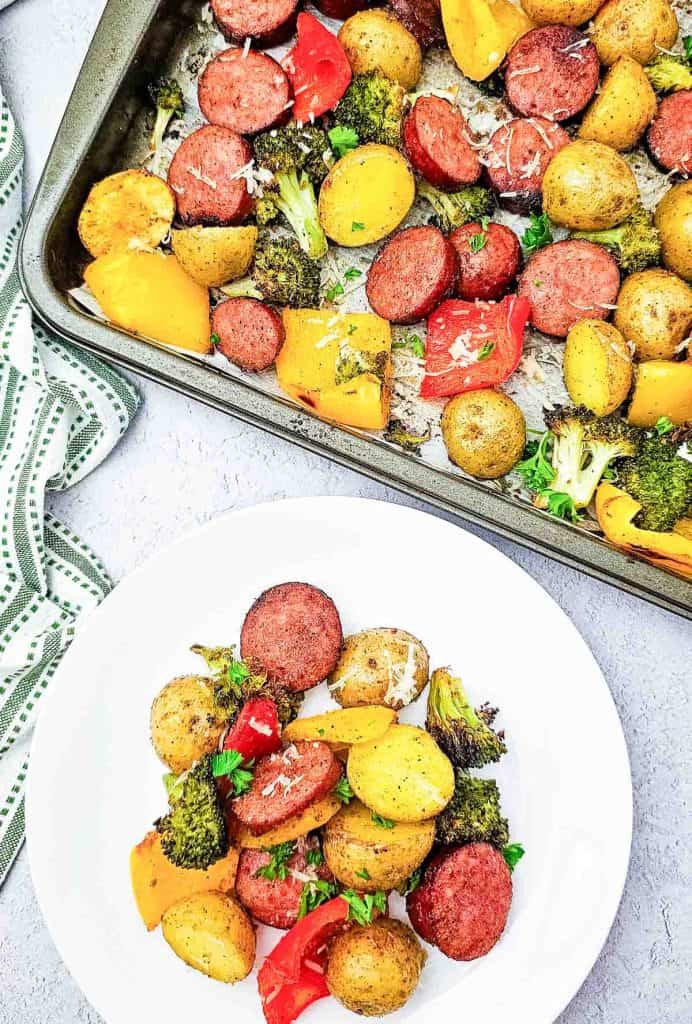 Roasted sausage and vegetables served on a white plate beside the baking sheet.