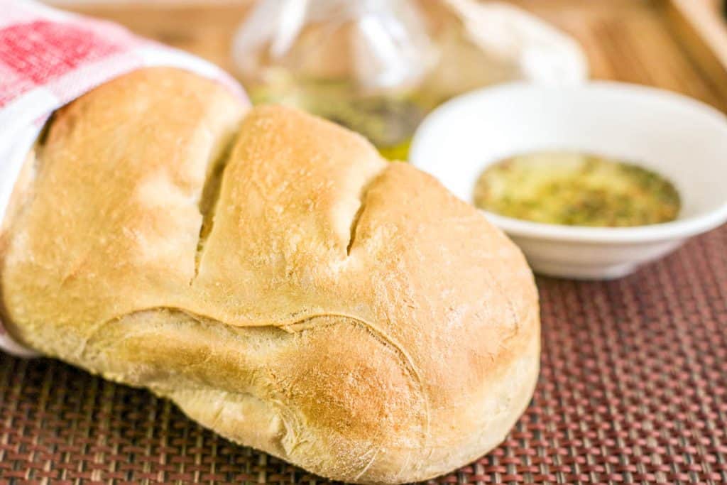 Freshly baked French bread loaf wrapped in a towel beside herb dipping oil.